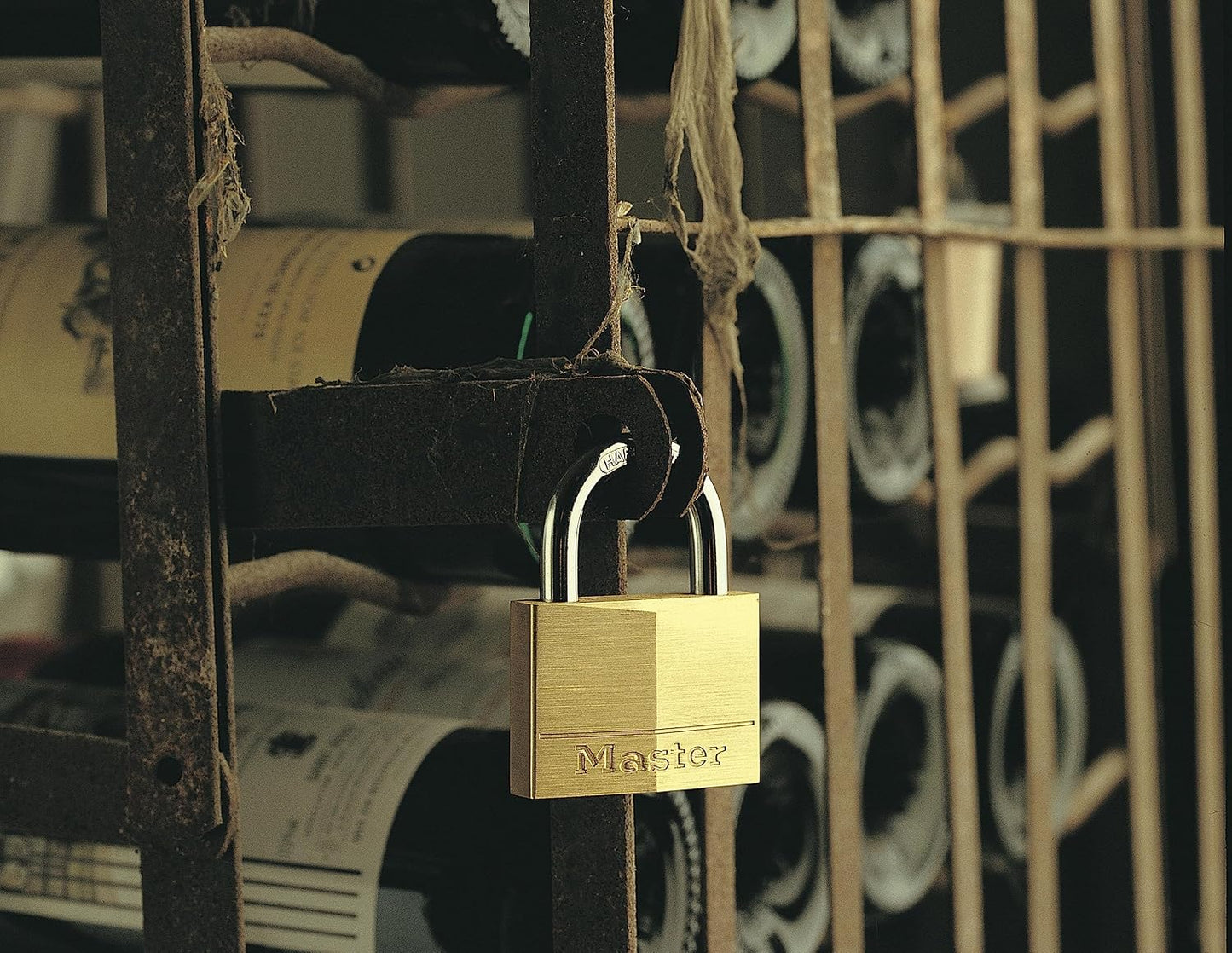 A brass Master padlock with silver shackle hooks hanging on a rustic black metal wine rack shelf, with wine bottles blurred in the background.